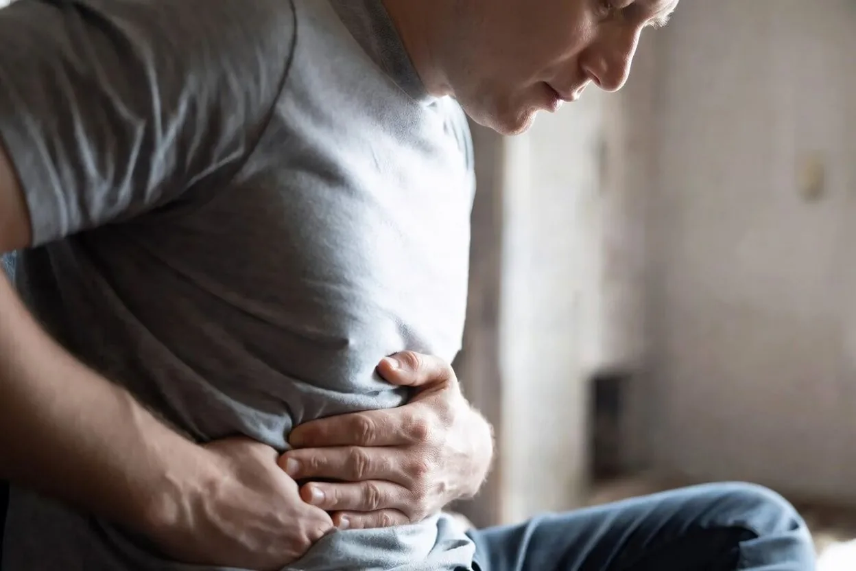 A person in a gray t-shirt sitting down and holding their abdomen in visible discomfort.